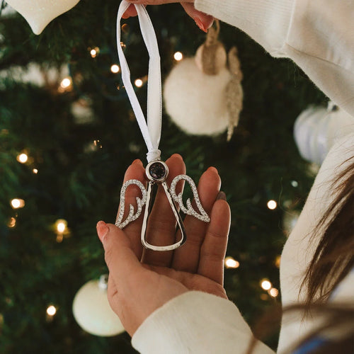 Acessorio em formato de anjo com foto personalizada no centro sendo segurado para pendurar na arvore de natal, com fita branca e fundo decorado com luzes e enfeites.
