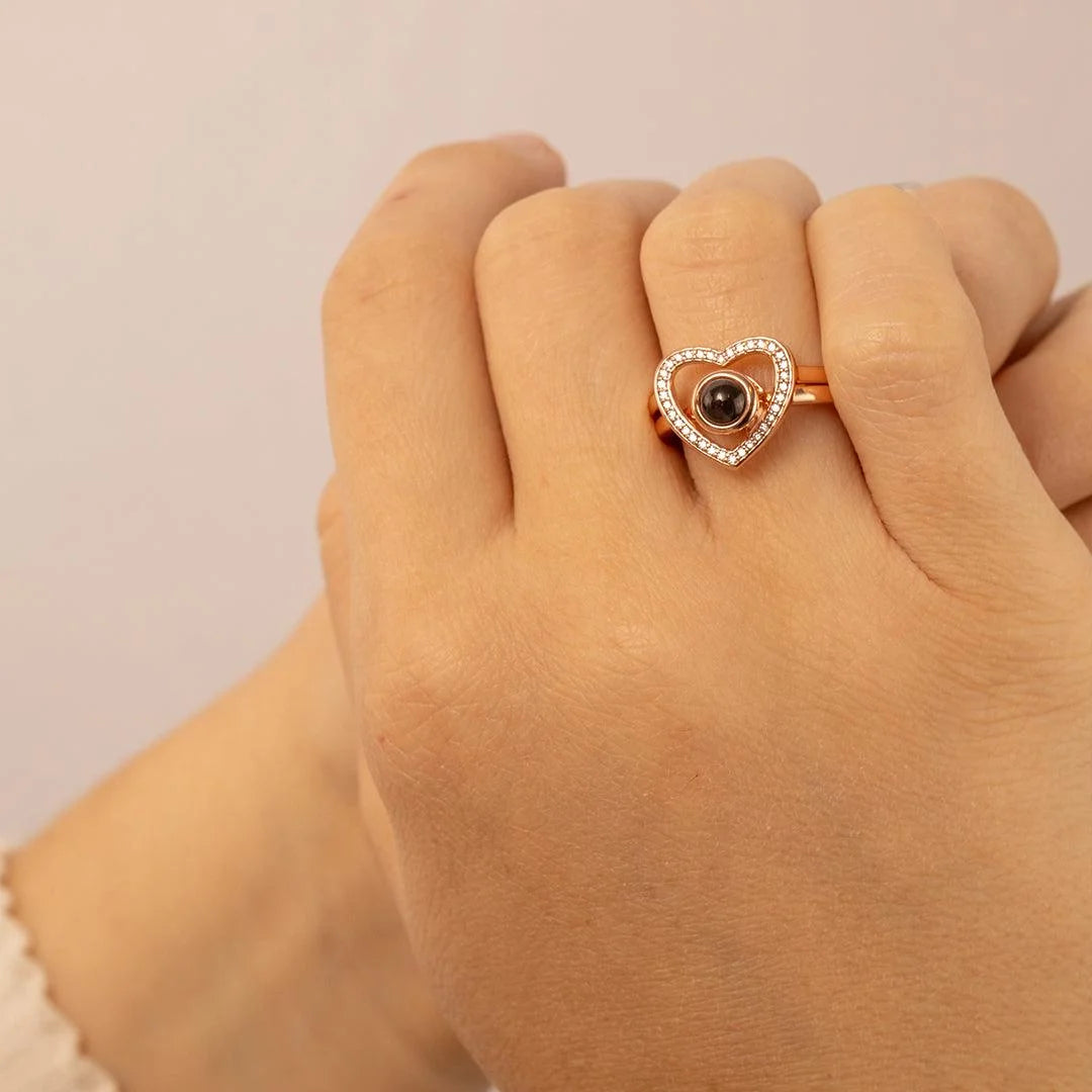 Anel rosé personalizado com coração cravejado usado na mão, com pedra central escura e fundo neutro.