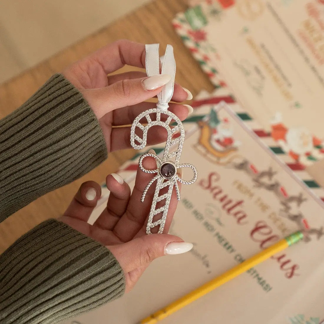 Enfeite de árvore de Natal em formato de bengala doce personalizada com foto, segurado sobre uma mesa com itens de presente ao fundo.
