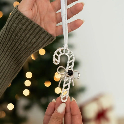 Enfeite natalino em formato de bengala doce personalizada sendo segurado pela mão, com árvore de Natal desfocada ao fundo.