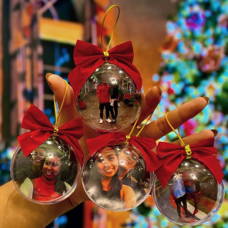 Conjunto de bolas de Natal personalizadas com fotos, exibidas na mão com laços vermelhos e fundo natalino desfocado.