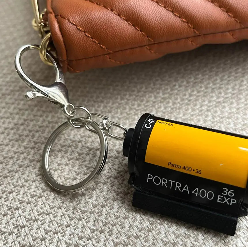 Chaveiro personalizado de câmera retrô com acabamento amarelo e preto apoiado sobre tecido, com argola metálica visível.