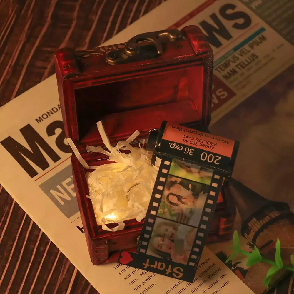 Chaveiro personalizado de câmera retrô dentro de caixa de presente com tira de filme fotográfico ao lado.