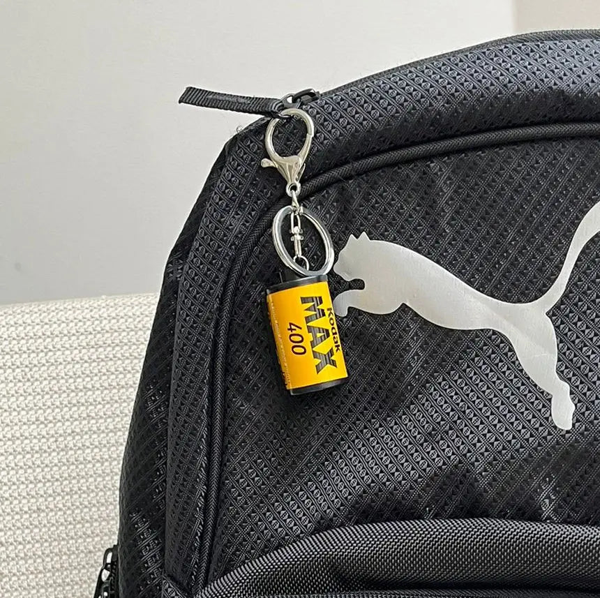 Chaveiro personalizado em formato de câmera retrô preso ao zíper de uma mochila preta.