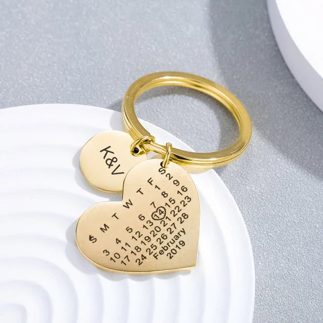 Chaveiro dourado personalizado com dois pingentes em formato de coração e círculo sobre fundo decorativo claro.