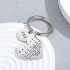Chaveiro prateado em formato de coração com gravação personalizada e plaquinha redonda com iniciais, sobre fundo claro decorativo.