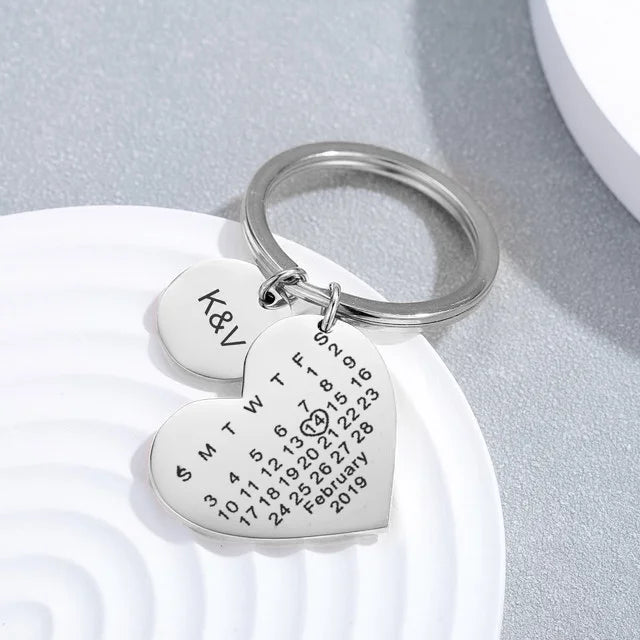 Chaveiro prateado em formato de coração com gravação personalizada e plaquinha redonda com iniciais, sobre fundo claro decorativo.
