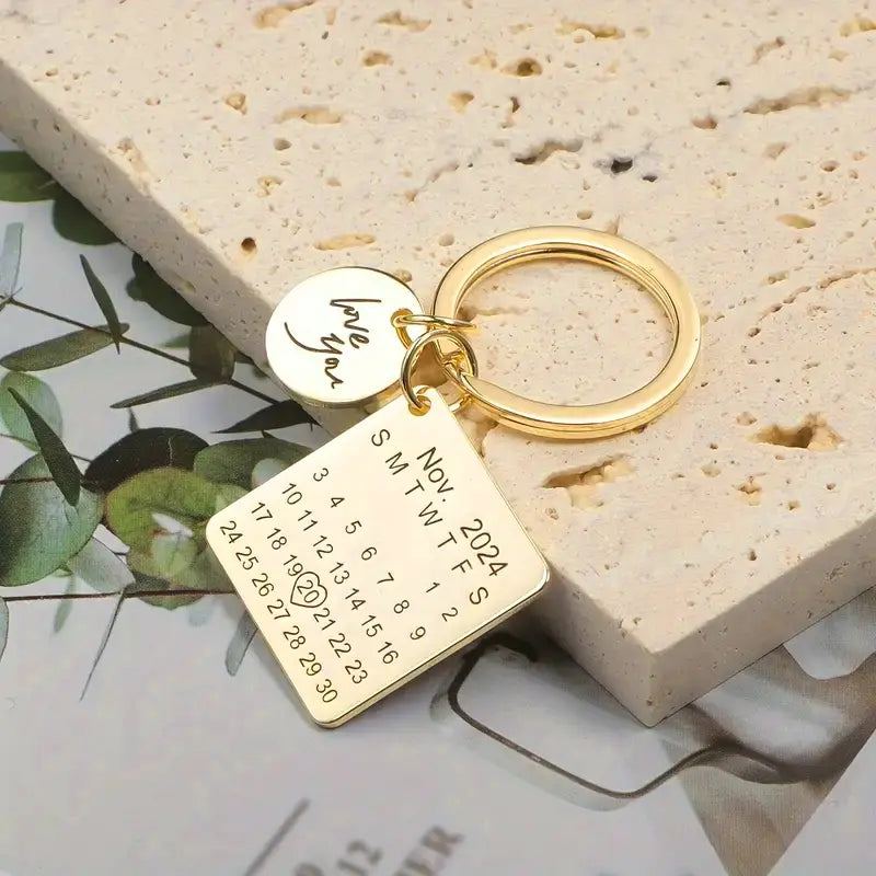 Chaveiro dourado personalizado com plaquinha quadrada exibindo calendário e disco com frase especial, sobre fundo decorativo.
