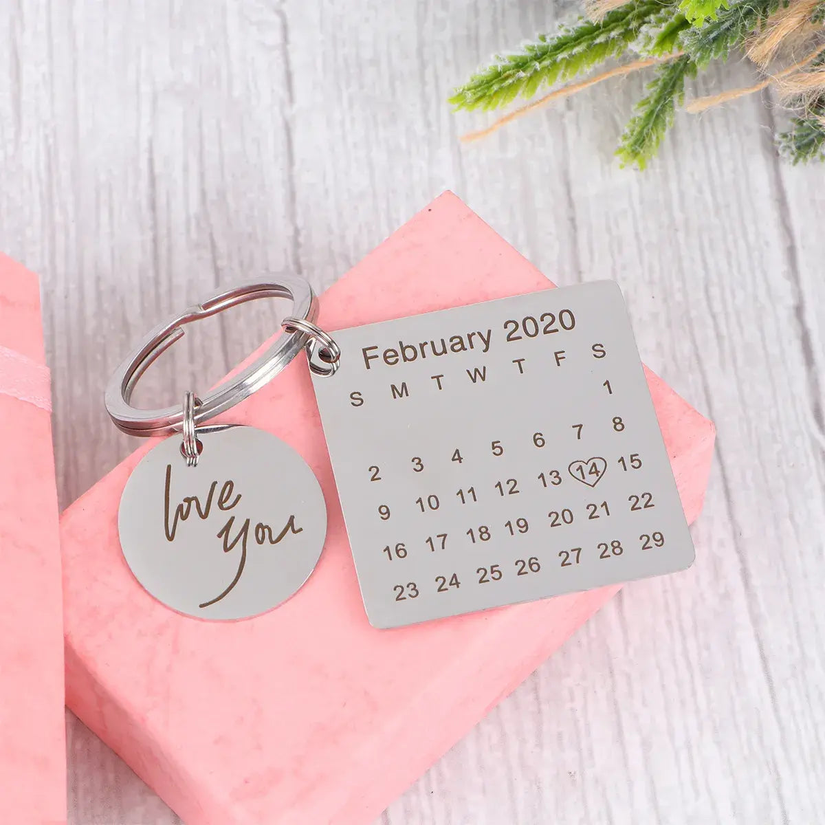 Chaveiro personalizado prateado com pingente redondo 'love you' e placa quadrada com calendário de fevereiro de 2020 apoiado sobre caixa rosa.