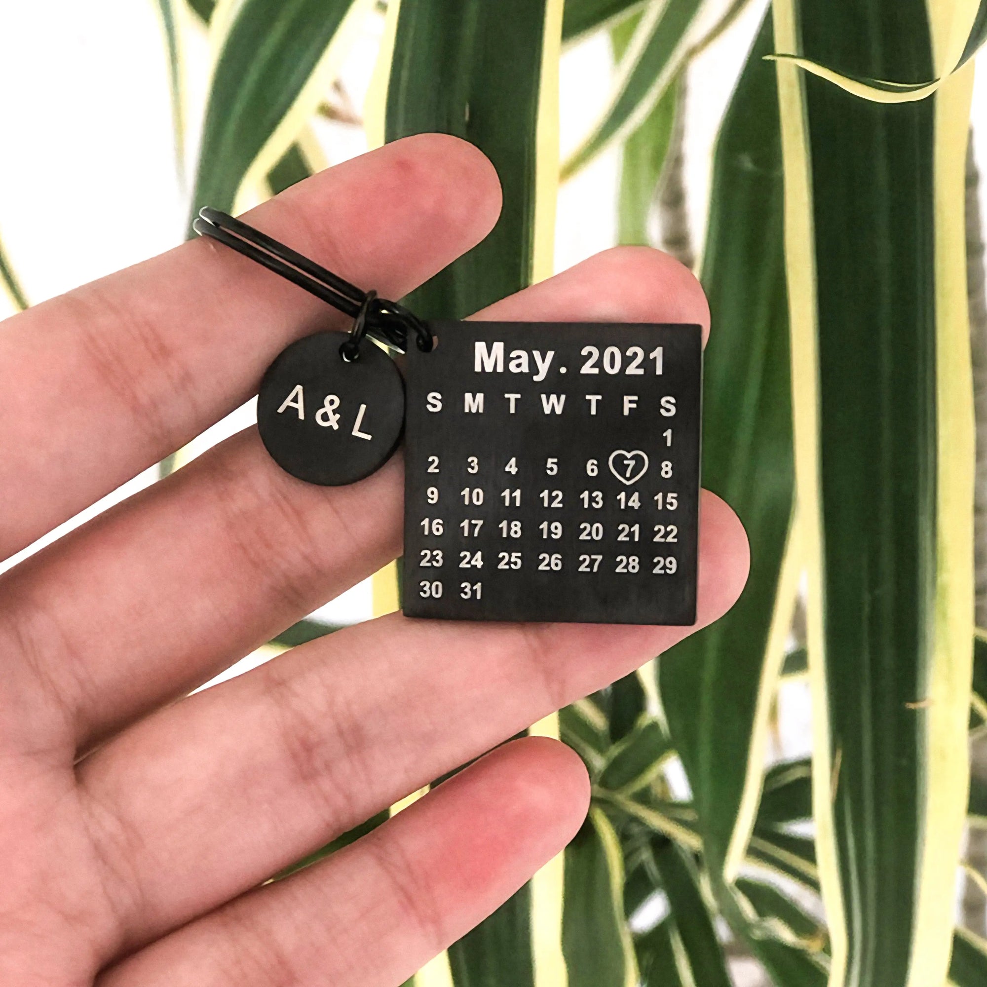 Chaveiro preto personalizado com iniciais e calendário, segurado na mão com fundo de folhas verdes.