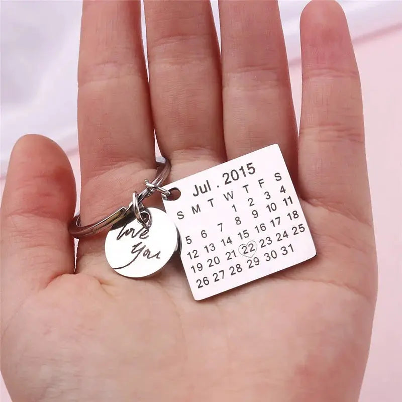 Chaveiro personalizado prateado com pingentes quadrado e redondo, exibido na mão com data e frase especial gravadas.