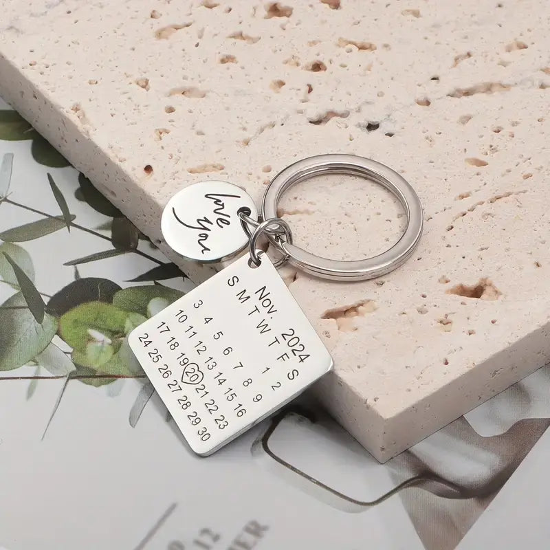 Chaveiro prata personalizado com placa quadrada de calendário e pingente circular sobre composição decorativa em fundo claro.
