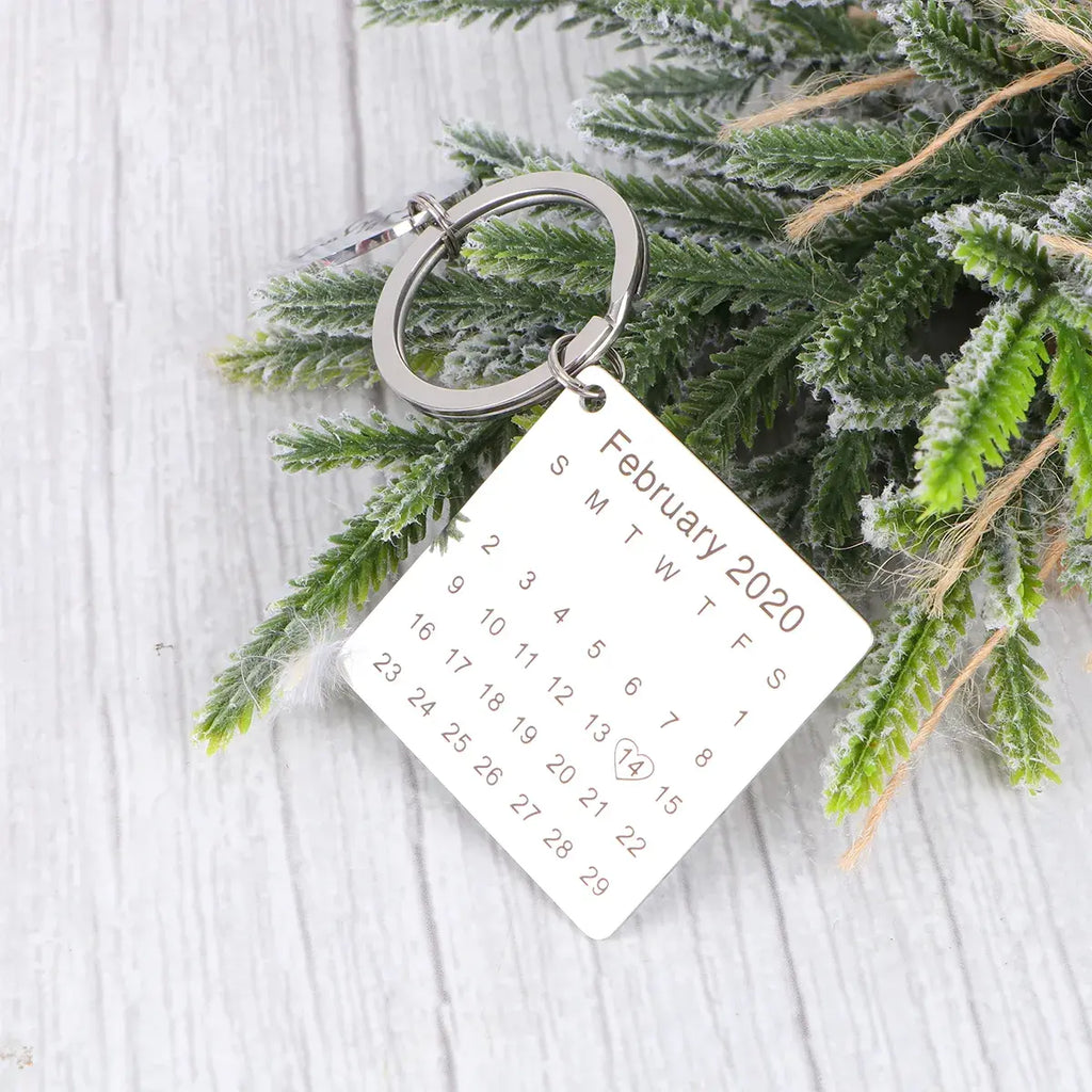Chaveiro personalizado de metal prateado com placa quadrada mostrando calendário de fevereiro de 2020 sobre fundo decorativo com folhagem.