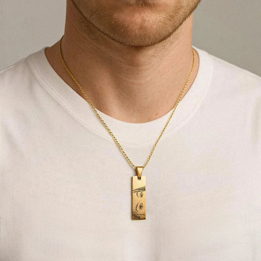 Colar dourado personalizado usado no pescoço de um homem, com pingente retangular com olhos femininos gravados no pingente pendente sobre camiseta branca.