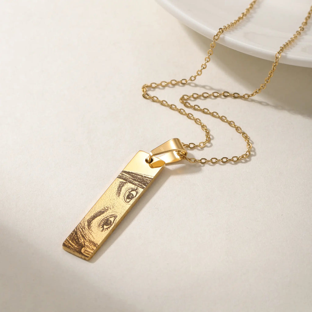 Colar dourado com pingente retangular gravado com olhos, em close sobre fundo claro.