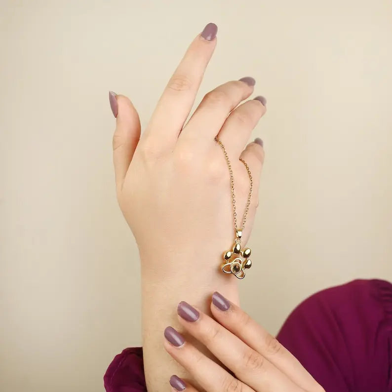 Colar dourado com pingente de patinha usado na mão, mostrando o detalhe delicado da corrente e do acabamento do produto.