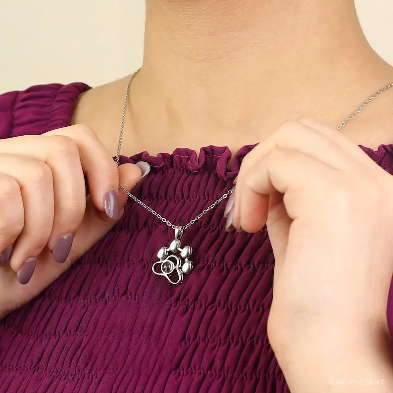 Colar de prata com pingente de patinha fofa sendo mostrado com as mãos sobre blusa roxa.