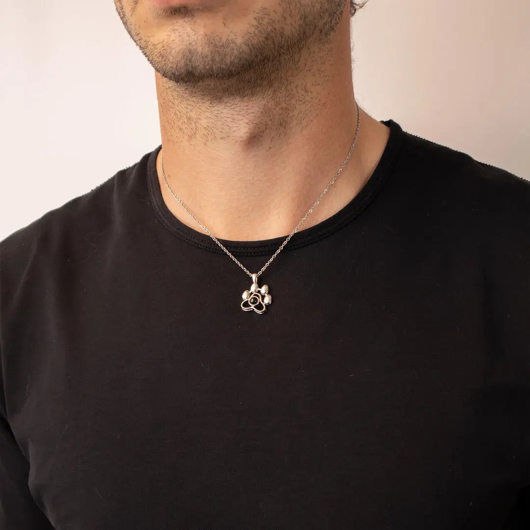 Colar de prata com pingente de patinha fofa usado no pescoço de homem sobre camiseta preta.