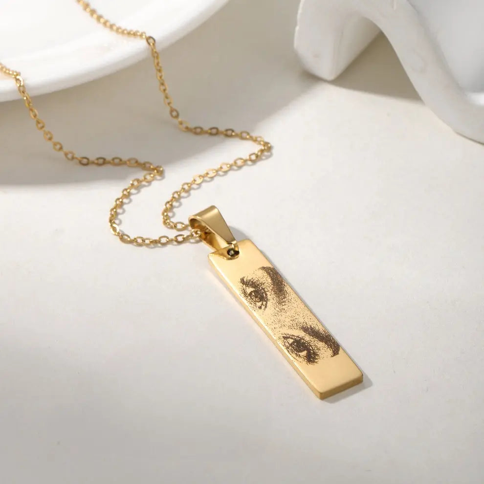 Colar personalizado dourado com pingente retangular e gravação artística de olhos, exibido sobre fundo neutro.