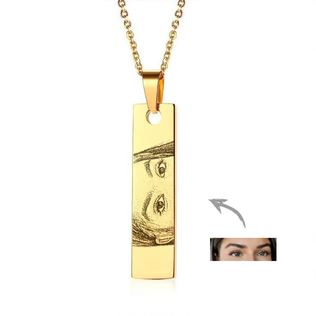 Colar personalizado dourado com pingente retangular gravado com desenho de olhos, em fundo branco, com detalhe do rosto aplicado à joia.