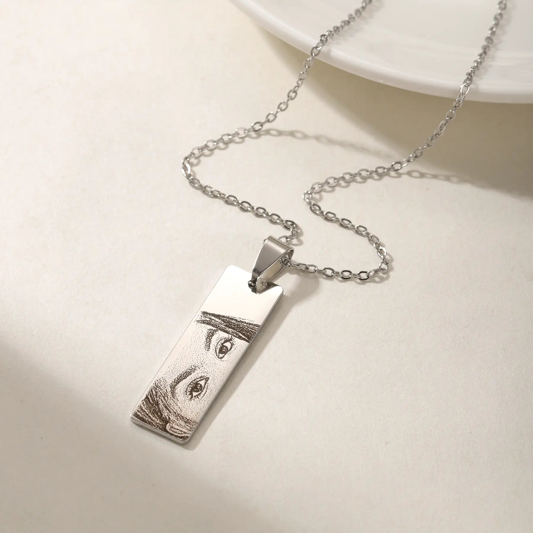 Colar personalizado de prata com pingente retangular gravado com desenho de olhos de uma mulher, em close mostrando qualidade da gravação sobre fundo claro.