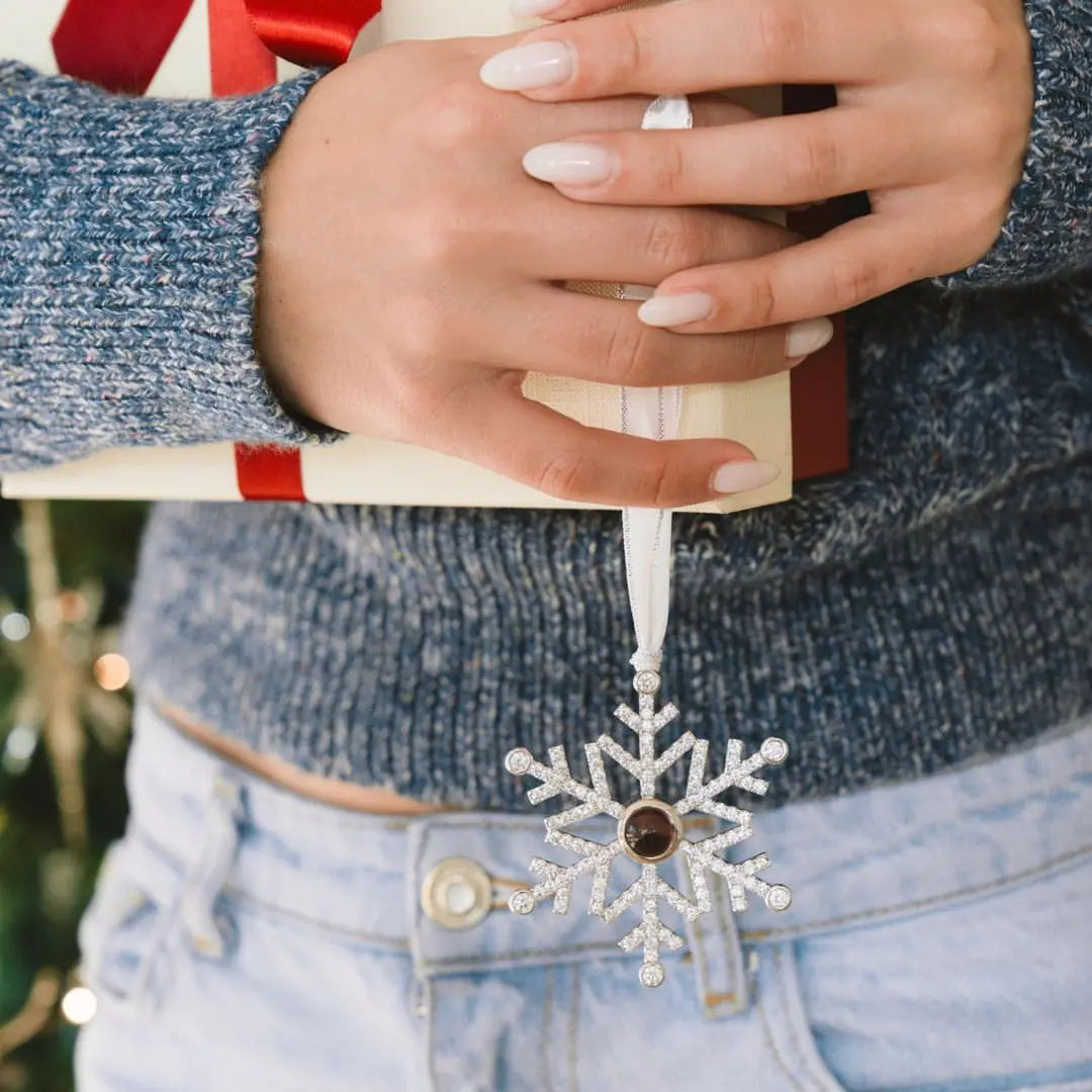 Acessório natalino em formato de floco de neve personalizado, segurado junto a um presente embrulhado com fita vermelha.