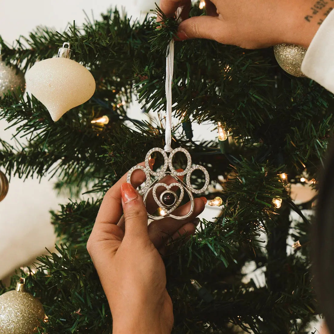 Enfeite de árvore de Natal em formato de pata de pet personalizado, sendo pendurado em um galho com decoração natalina ao fundo.