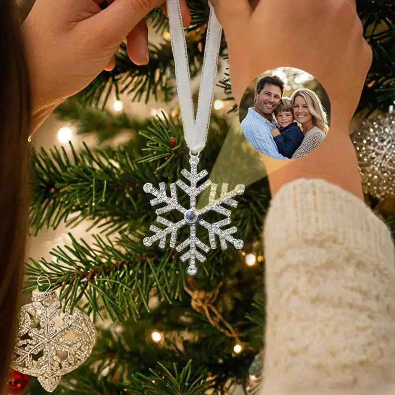 Acessório para árvore de Natal em formato de floco de neve com foto personalizada de família no centro como exemplo, pendurado em frente aos galhos decorados da árvore.